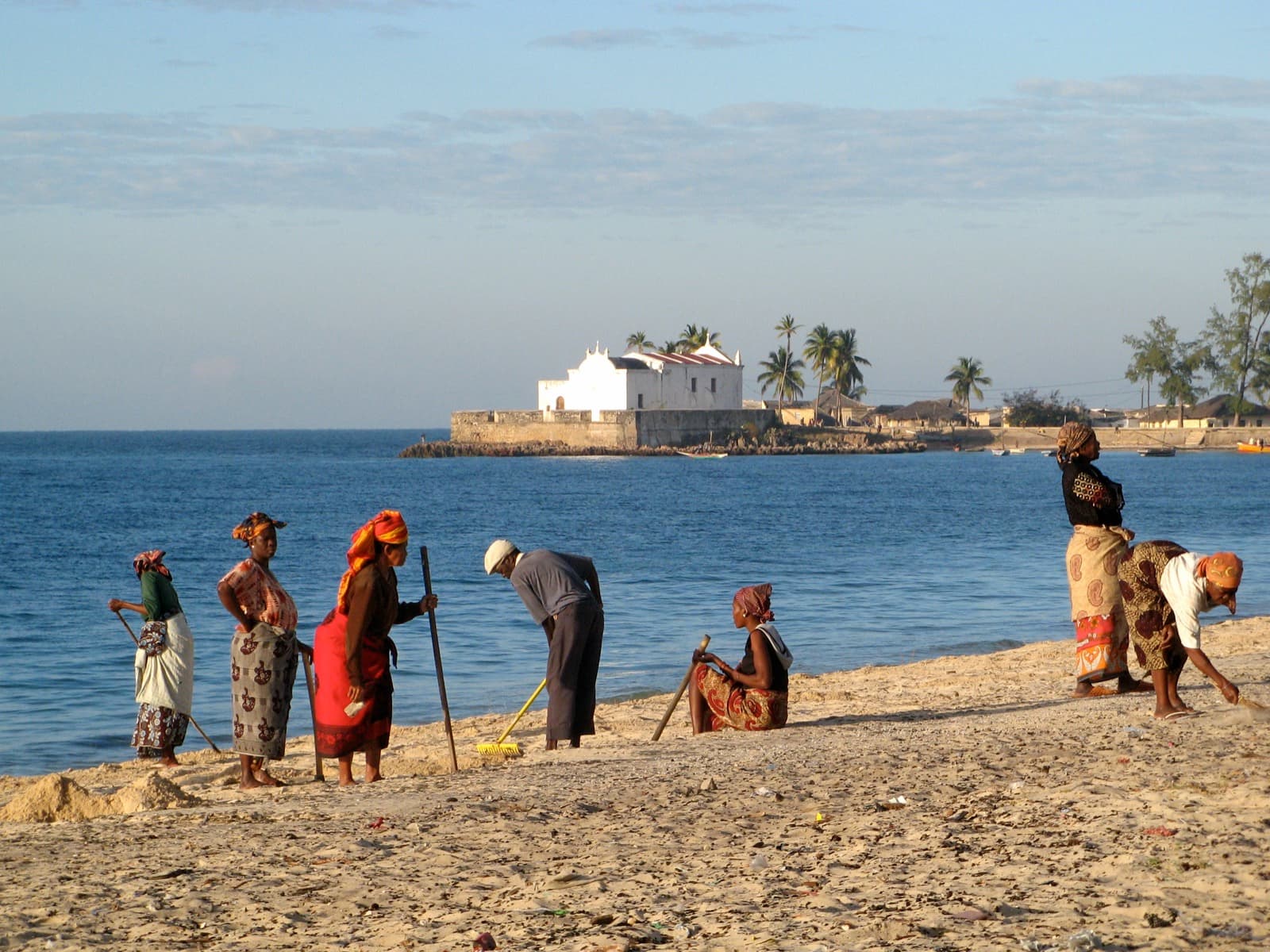 Pessoas limpando uma praia em Moçambique, com uma igreja branca e palmeiras ao fundo, à beira do mar